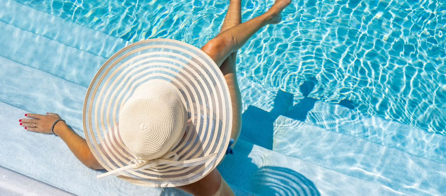 Top view of a woman wearing an elegant beige sun hat, sitting cross-legged in the water on the steps of a stone-floored pool with translucent water.