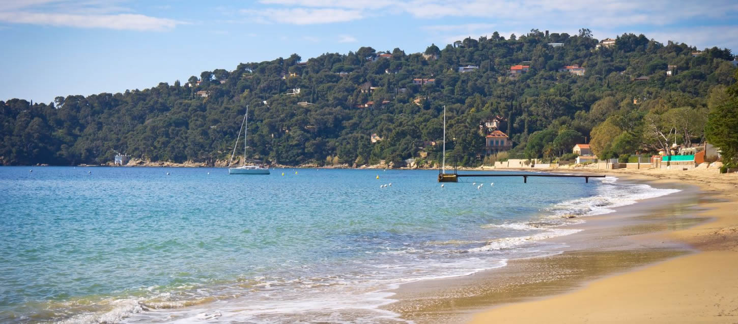 View of Tahiti Beach with its turquoise water, fine sand, a wooden pier with a sailboat at the end, the pine-covered hill with the roofs of houses, and other houses near the beach.