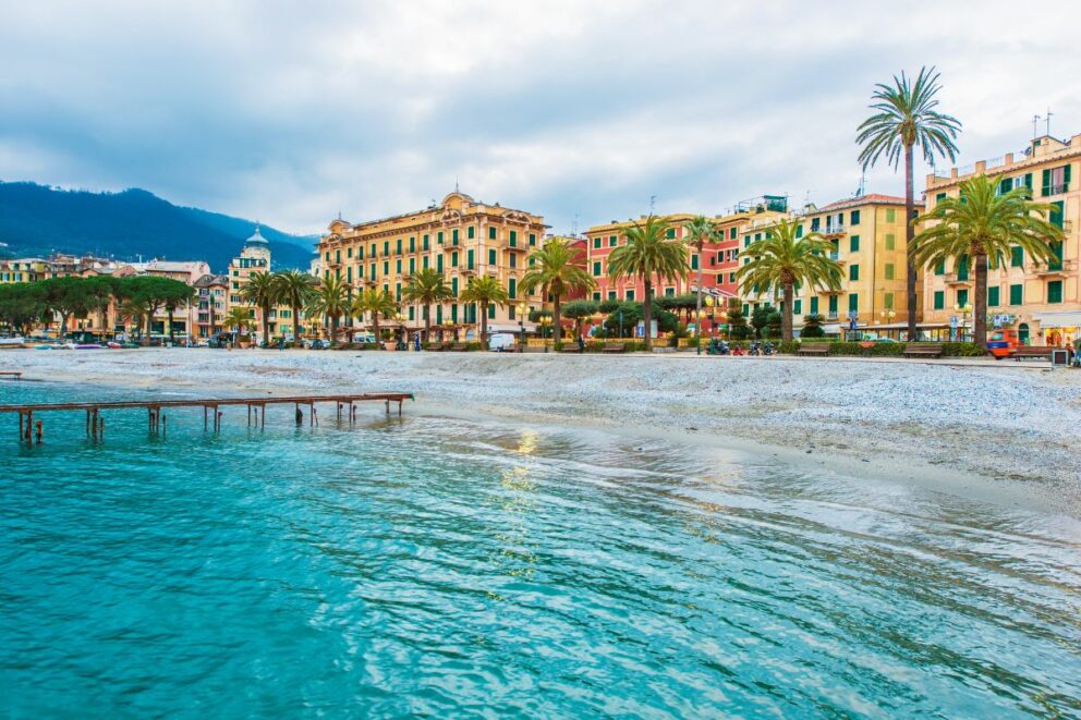 Colorful seaside buildings and palm trees line the waterfront of Santa Margherita Ligure on the Italian Riviera, with turquoise water and distant green hills under a cloudy sky.