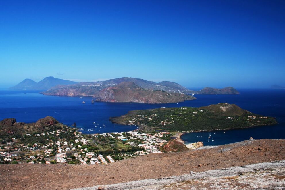 A panoramic view of the Aeolian Islands, with the town of Vulcano in the foreground and the island of Lipari visible in the distance across the blue sea. The sky is clear, and several boats are anchored in the bay.