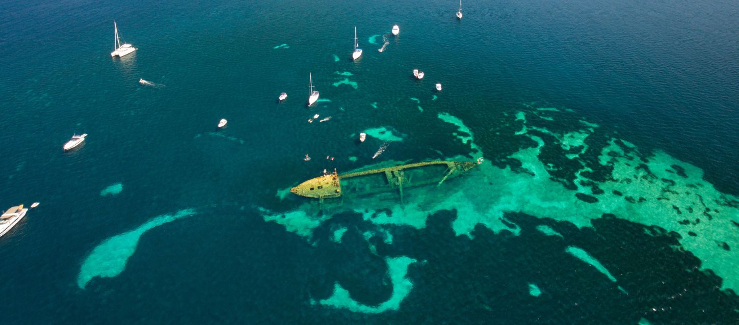 Aerial view of a shipwreck submerged in clear turquoise waters near Dugi Otok, Croatia, surrounded by small boats and yachts floating above the vibrant coral seabed.