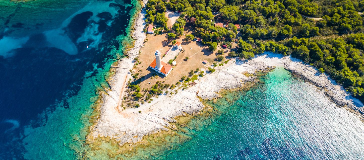 Aerial view of the Veli Rat lighthouse on Dugi Otok Island, Croatia, surrounded by turquoise waters and dense pine forest, with rocky white shores and crystal-clear sea under a bright blue sky.