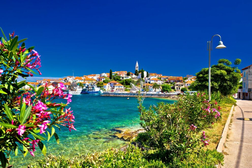 Scenic coastal view of Kali village near Zadar, Croatia, showing turquoise water, fishing boats in the harbor, colorful houses with orange rooftops, and vibrant pink flowers in the foreground under a deep blue sky.