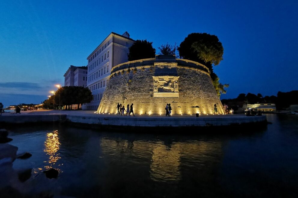 Evening view of Zadar Old Town, Croatia, showing the illuminated stone city walls and a carved gate reflected on calm water, with people walking nearby and historic buildings under a deep blue twilight sky.