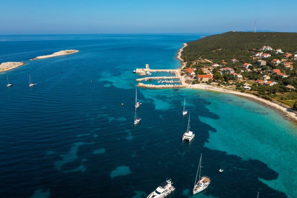Aerial view of sailboats and yachts anchored in turquoise waters near the island of Premuda, Croatia, with a small marina and coastal village surrounded by green hills and clear blue sea.