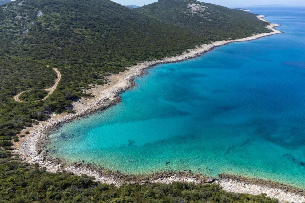 Aerial view of a rocky coastline on the island of Molat, Croatia, with turquoise waters, lush green hills, and a narrow dirt path winding along the shore.