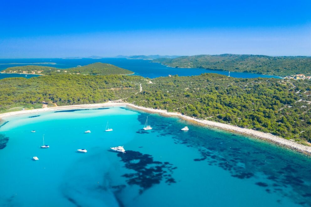 Aerial view of Sakarun Beach on Dugi Otok Island, Croatia, featuring turquoise shallow waters, anchored yachts, and lush green hills under a clear blue sky.