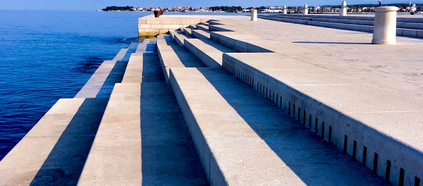 View of the Sea Organ in Zadar, Croatia — a series of wide marble steps descending into the Adriatic Sea, with slots that produce musical sounds from the movement of the waves, under clear blue skies.