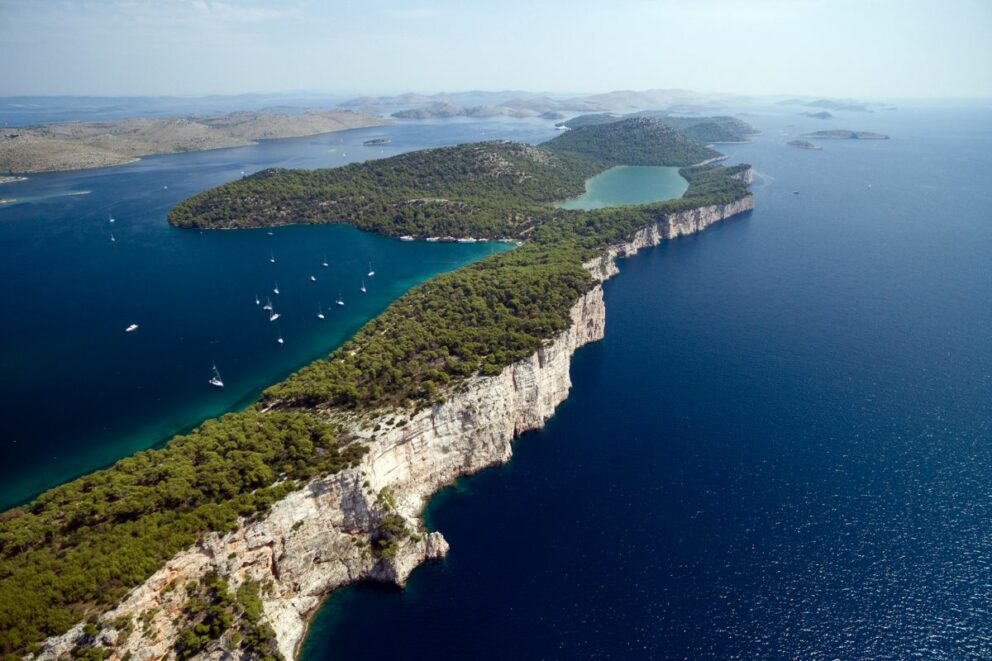 Aerial view of Telašćica Nature Park in Croatia, showing dramatic white cliffs dropping into the deep blue Adriatic Sea, green forests, and a turquoise saltwater lake surrounded by anchored sailboats.