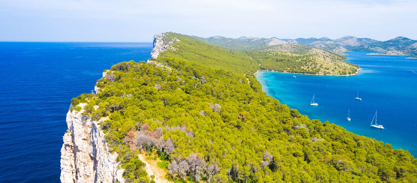 Aerial view of Telašćica Nature Park on Dugi Otok Island, Croatia, showing steep white cliffs, lush green forests, turquoise bays, and sailboats anchored in calm Adriatic waters under a clear sky.