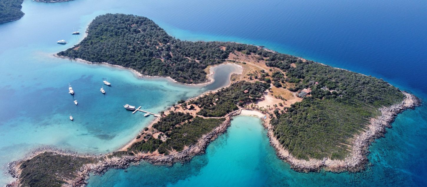 Aerial photograph of Sedir Island, also known as Cleopatra Island, located in the Gulf of Gökova, Turkey. The island is covered in lush green vegetation and surrounded by clear turquoise and deep blue water. A small, sandy beach, is visible on the island's shore. Several boats are anchored in the calm waters near a pier, suggesting it is a popular spot for visitors. The island has a distinctive shape with a central inlet of water