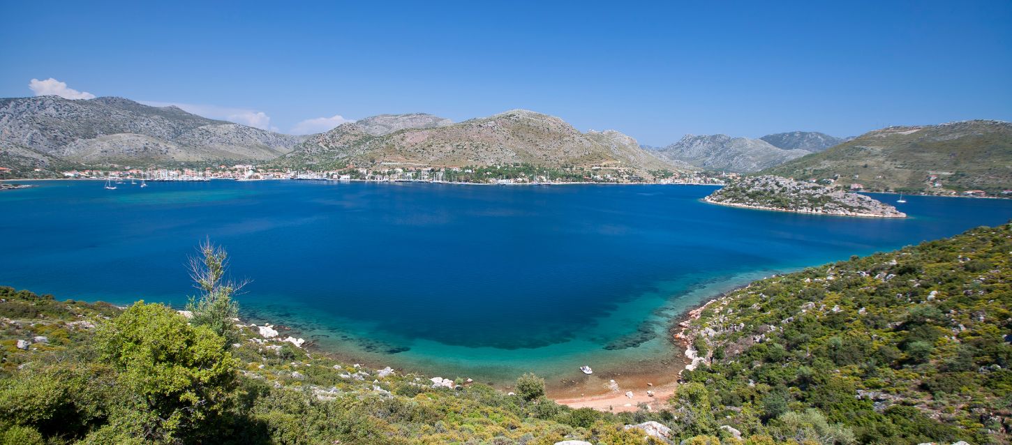 This is a scenic photograph of a bay surrounded by hills and a small town. The water in the bay is a deep blue, with a lighter turquoise color near the shore, suggesting shallow water. In the background, there are mountains and hills covered in green vegetation, with a town built along the waterfront. The sky is clear and bright blue. The foreground features a rocky shoreline with green bushes and a small, sandy beach
