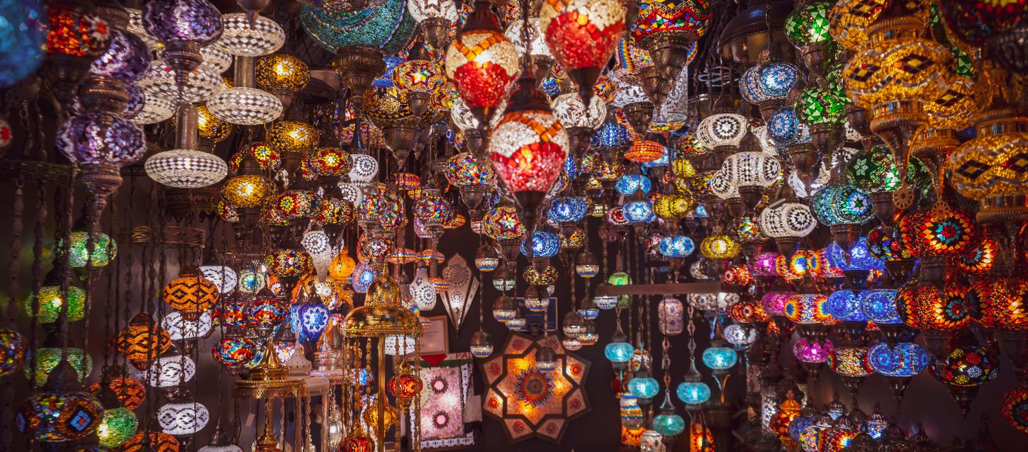 Large collection of colorful, ornate lamps and lanterns hanging from a ceiling. The lamps are made of mosaic glass in various colors like red, blue, green, and orange, and they are suspended at different heights, The lighting from the lamps illuminates the scene, highlighting the intricate patterns and designs of each piece