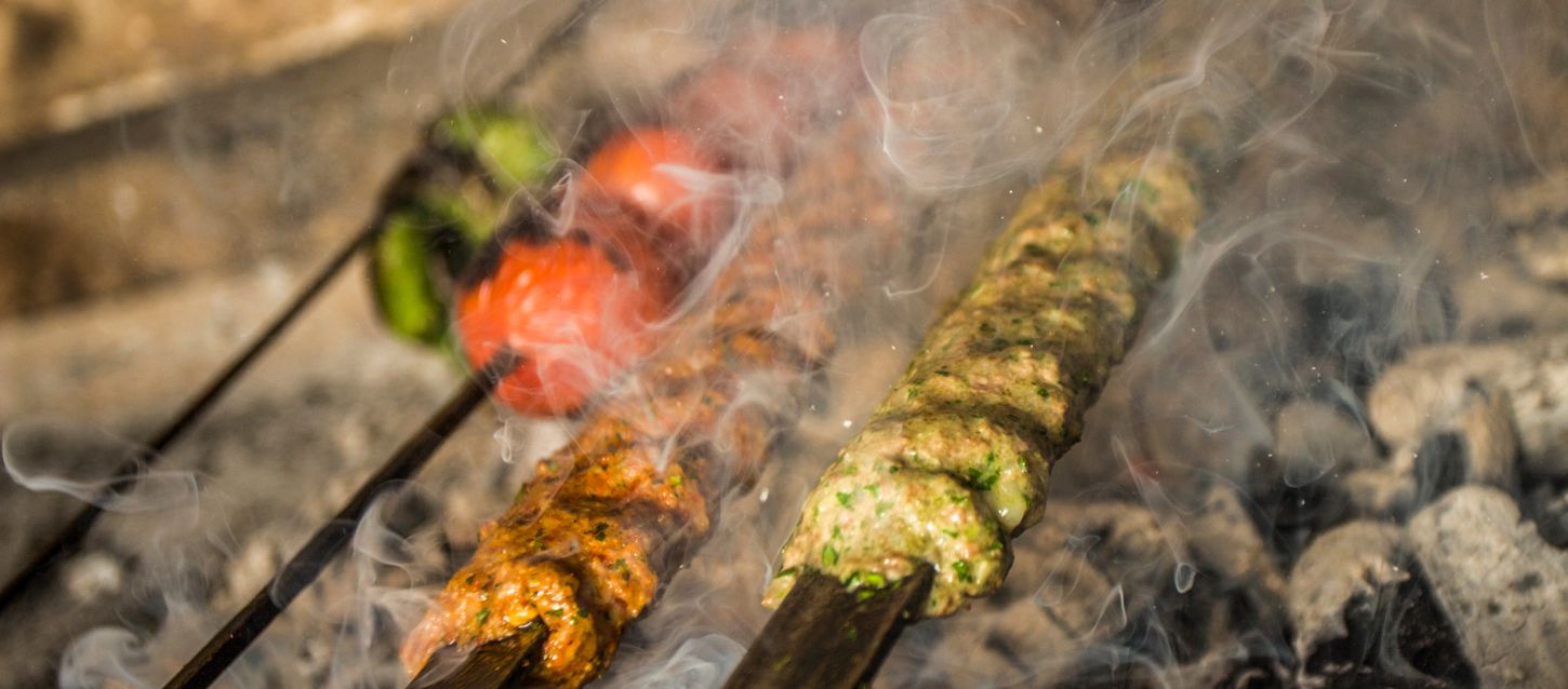 The image shows two skewers of meat and vegetables cooking over charcoal. The skewers are smoking, and the meat appears to be a type of tikka, possibly chicken, marinated in spices. One skewer has a reddish-orange marinade, while the other has a greenish-brown marinade. There are also what appear to be tomatoes and a green pepper on a separate skewer in the background. The cooking process is creating a lot of smoke, which is visible throughout the image. The meat is cooking directly on the grill, suggesting it is being roasted over a fire