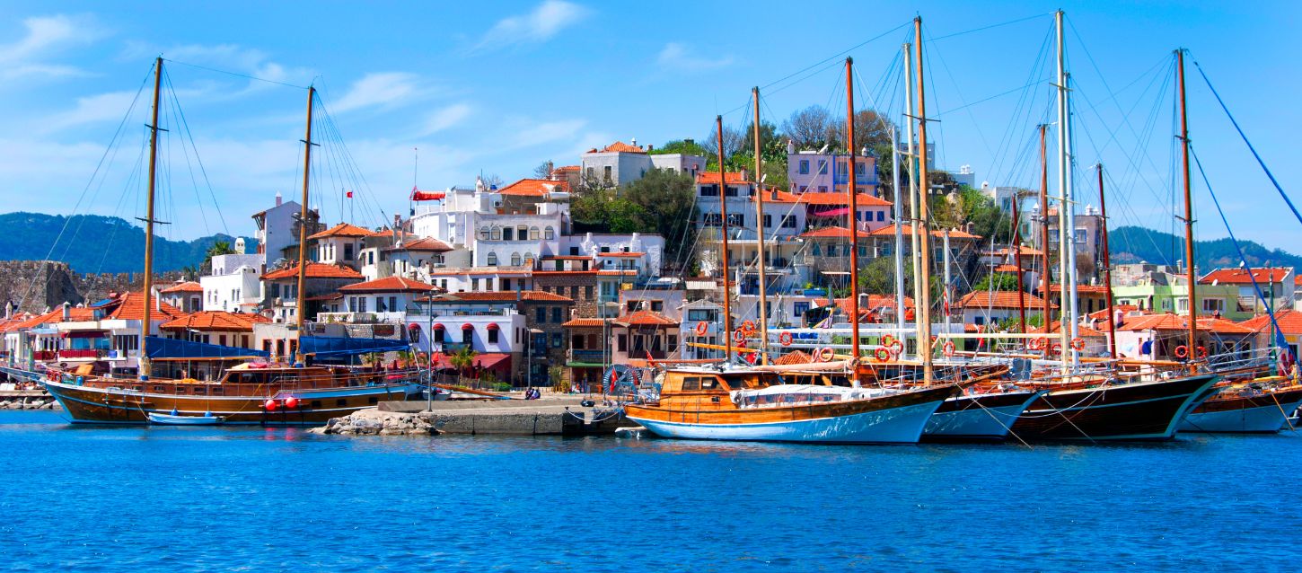 This is a photograph of a harbor in what appears to be a Mediterranean or Aegean town, possibly Marmaris, Turkey. The foreground shows several traditional wooden sailing boats, known as gulets, docked in a marina with calm, blue water. In the background, a hillside is densely populated with white buildings featuring red-tiled roofs. The sky is clear and blue, and distant green hills or mountains are visible on the horizon.