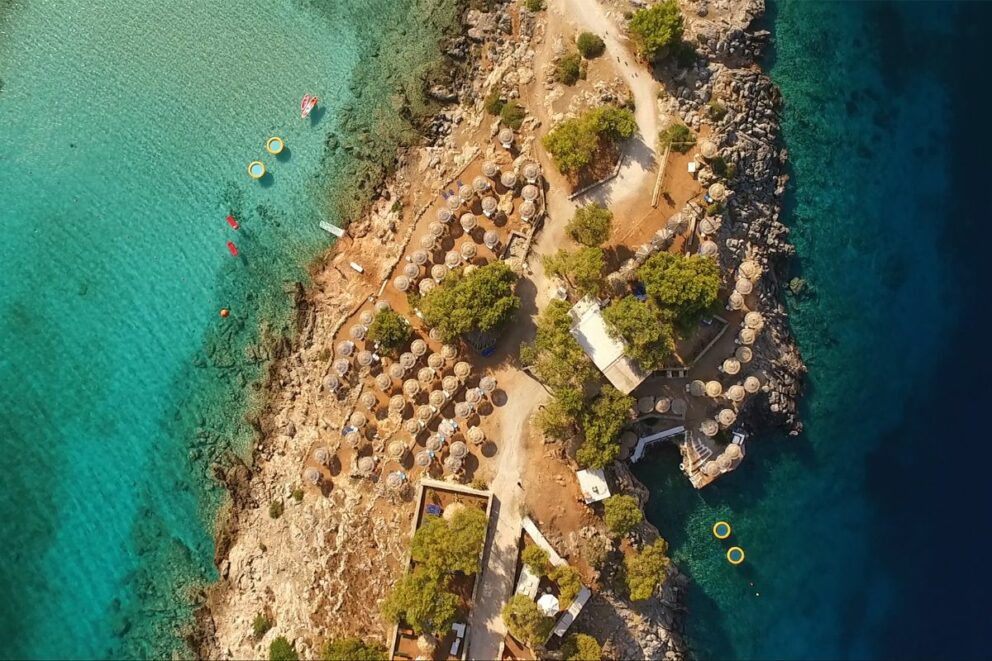 Aerial view of a rocky beach resort with many straw umbrellas, sunbeds, and clear turquoise water.