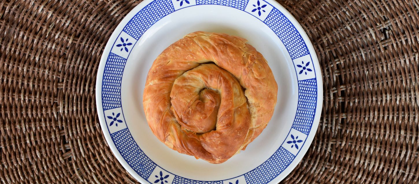 A spiral-shaped Tyropita (Greek cheese pie) on a white plate with blue patterns, set against a woven placemat