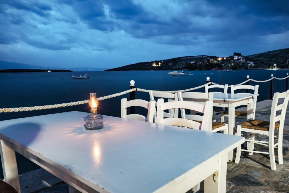 A seaside restaurant table with white chairs and a lit lantern at dusk, overlooking the water towards the village of Amaliapoli, Greece.