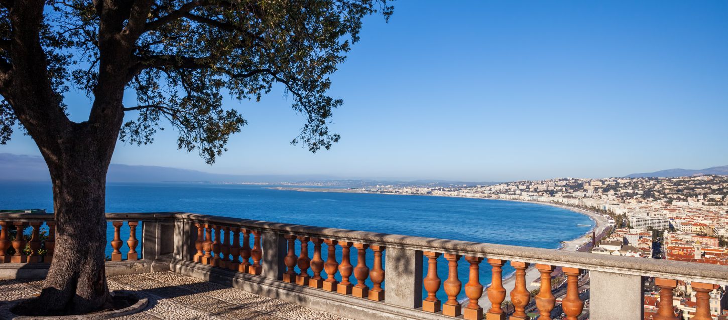 A walkway lined with lush vegetation leads to a viewpoint with a red railing overlooking the Baie des Anges. From there, the view opens to the coastline and the turquoise waters of the Mediterranean Sea, shimmering under the sunlight