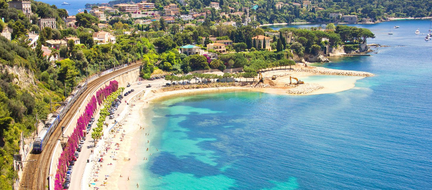 View of Cap Ferrat with its luxury villas surrounded by pine trees, facing its white sand beach and turquoise waters with yachts anchored offshore.