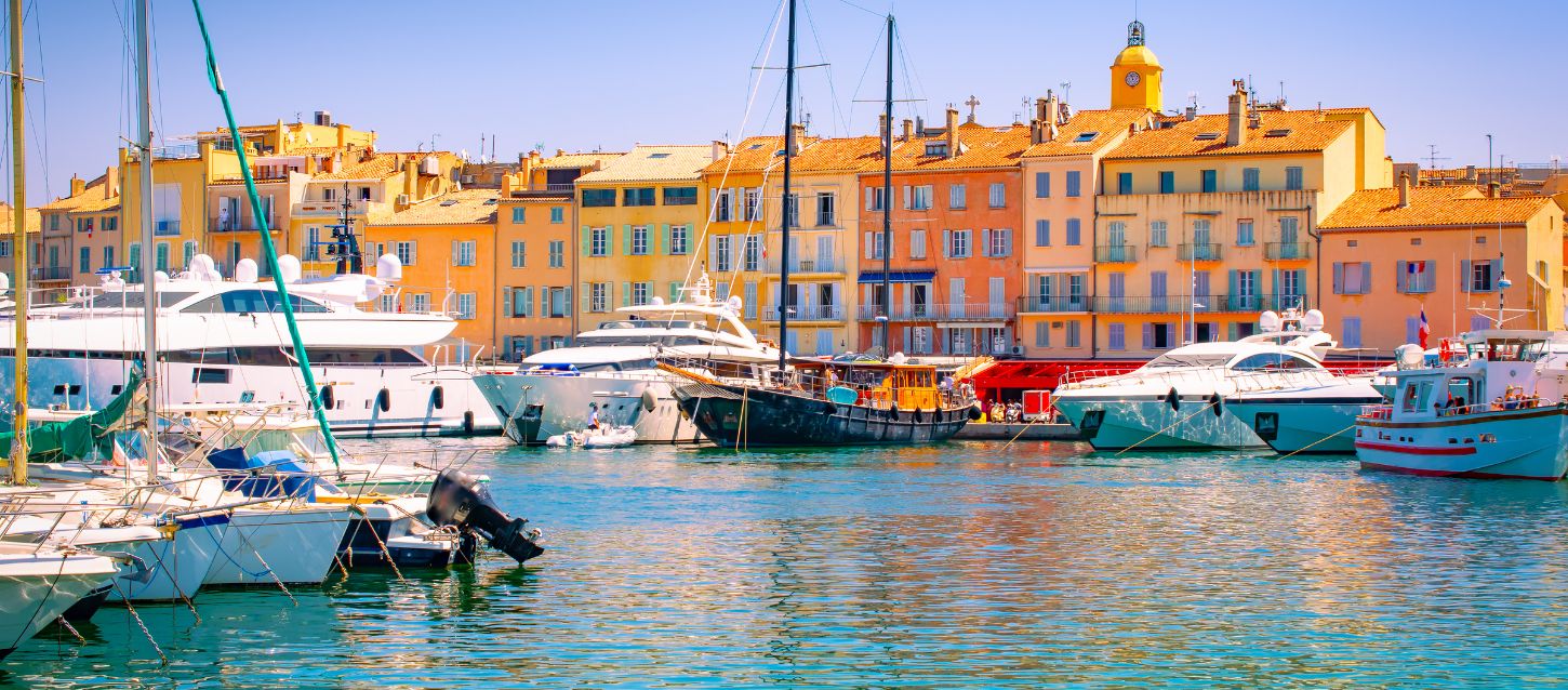 Port of Saint Tropez with luxury sailboats and yachts moored, classic sailboat, town facades with their typical golden-toned buildings, sunlit tiled roofs, antique wooden shutters in light tones, and bell tower under a clear sky.