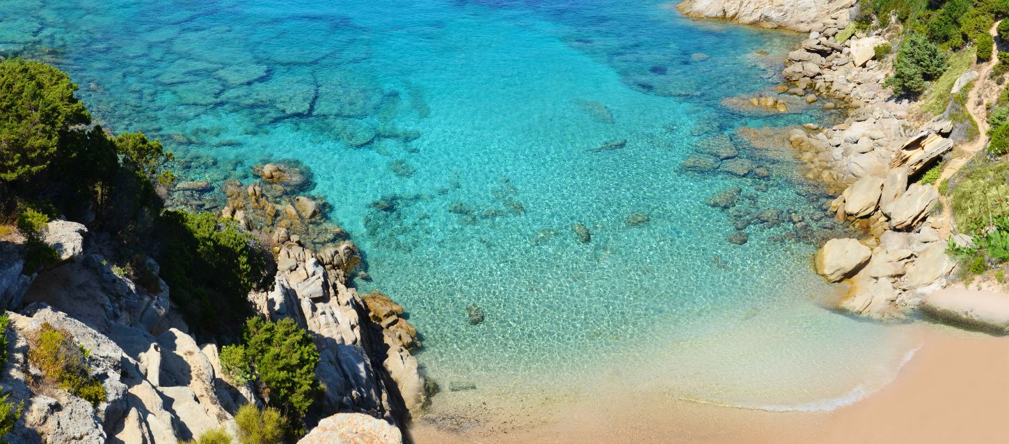 Cala de L'escalet with crystal clear turquoise waters, clean seabed with some rocks, fine golden sand, smooth rocks covered with lush green vegetation, nature trail