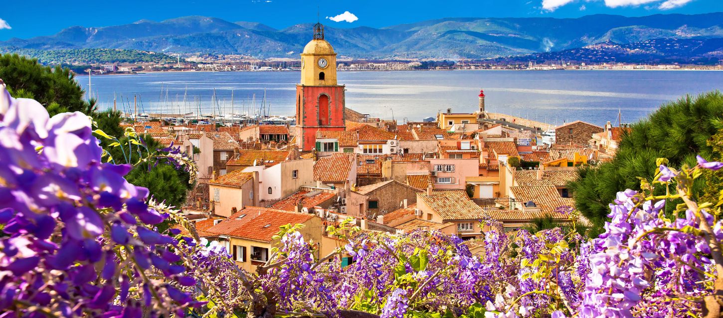 View of the coastal town of Saint Tropez with terracotta rooftops and the yellow clock tower overlooking the harbour, framed by vibrant purple flowers in the foreground and mountains in the background with the sea