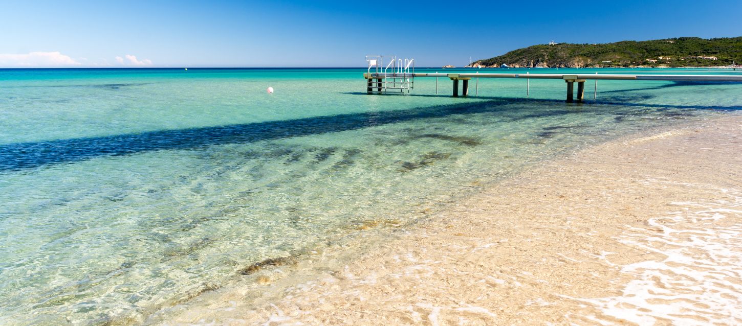 View of Pampelonne Beach with its golden sand and crystal clear waters, calm sea in different shades of turquoise. Swimming platform with ladder for easy access to the water. Coastline with trees and cliffs, yachts visible in the distance.