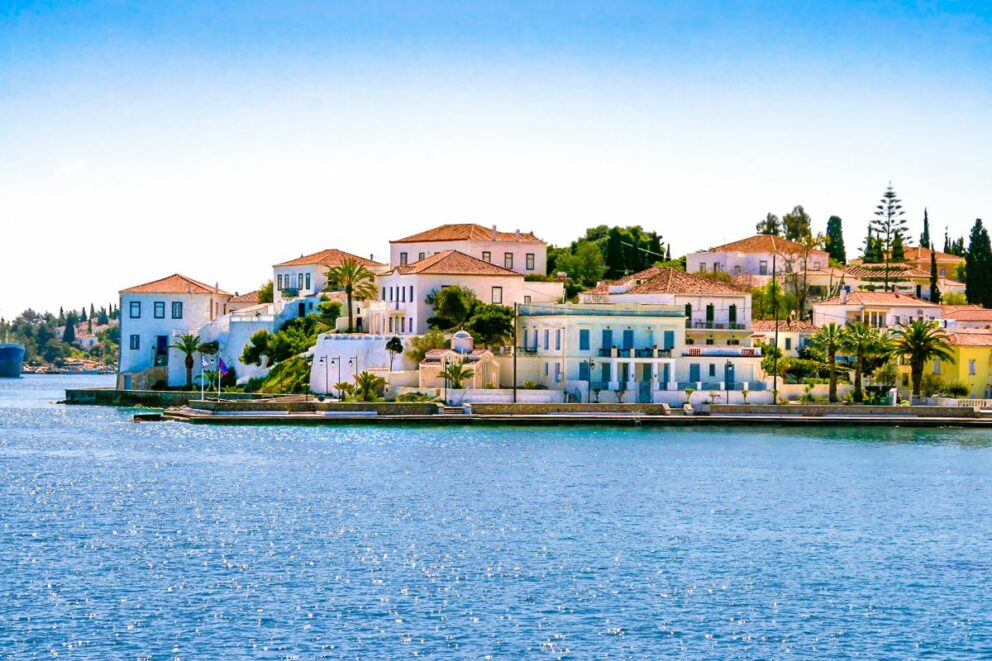 A view of the coastal town of Spetses, Greece, featuring traditional white buildings with red-tiled roofs along the waterfront.