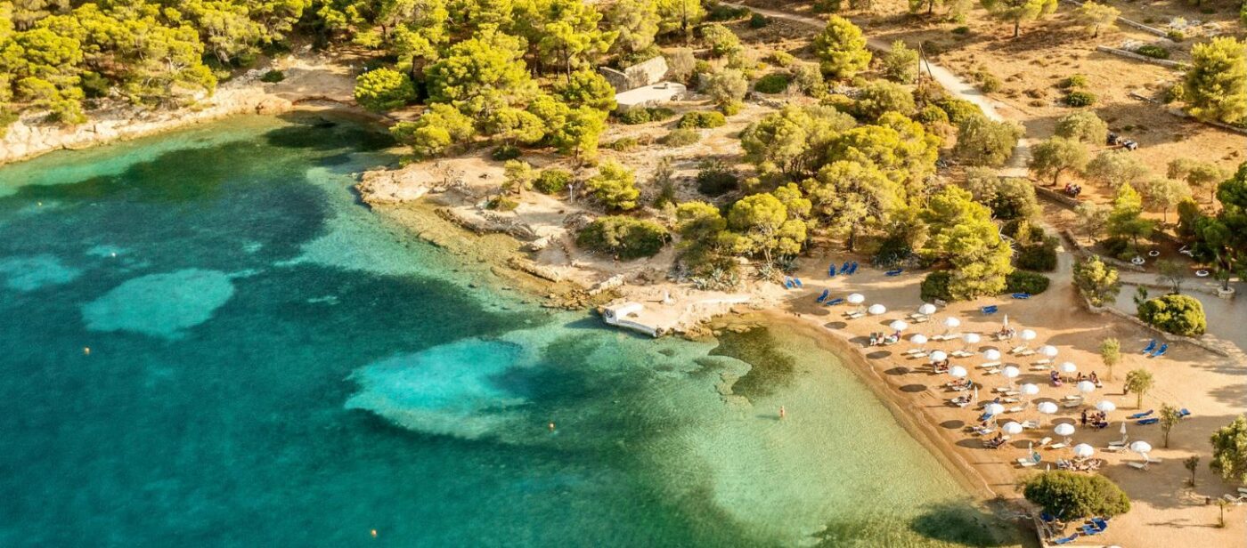 An aerial view of Zogeria Beach in Spetses, featuring clear turquoise water, a sandy shore with white umbrellas and sun loungers, and a lush green pine forest hillside.