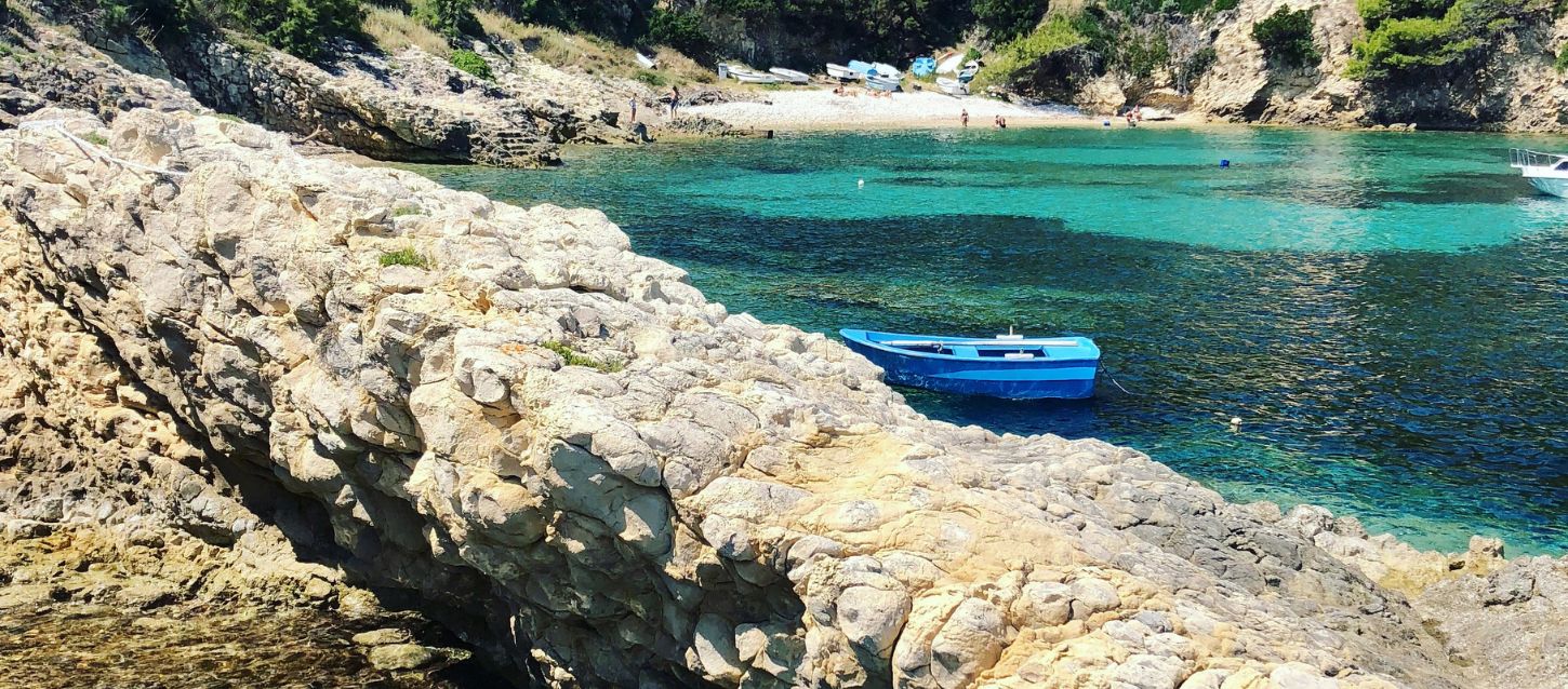 rocky coastline with a small, secluded beach in the background. The water is a striking, clear turquoise color. In the foreground, a large, jagged rock formation extends into the water from the left. A small, bright blue boat is anchored in the calm water between the rocks and the beach. In the background, a few people are visible on the sandy shore, and several other boats are pulled up onto the sand