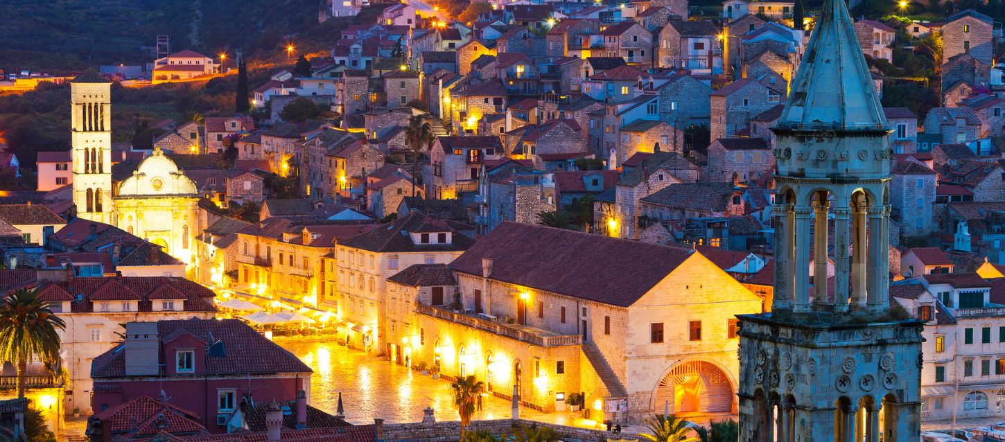 nighttime photograph of a coastal town, likely Hvar, Croatia. The town is built on a hillside, with many buildings illuminated by warm lights. In the foreground, there are two prominent bell towers and a large building with arched entrances, possibly the Hvar Arsenal. The lights from the buildings reflect on the water in the foreground, creating a bright, glowing effect. The sky is a deep blue, suggesting it is either dusk or early night.