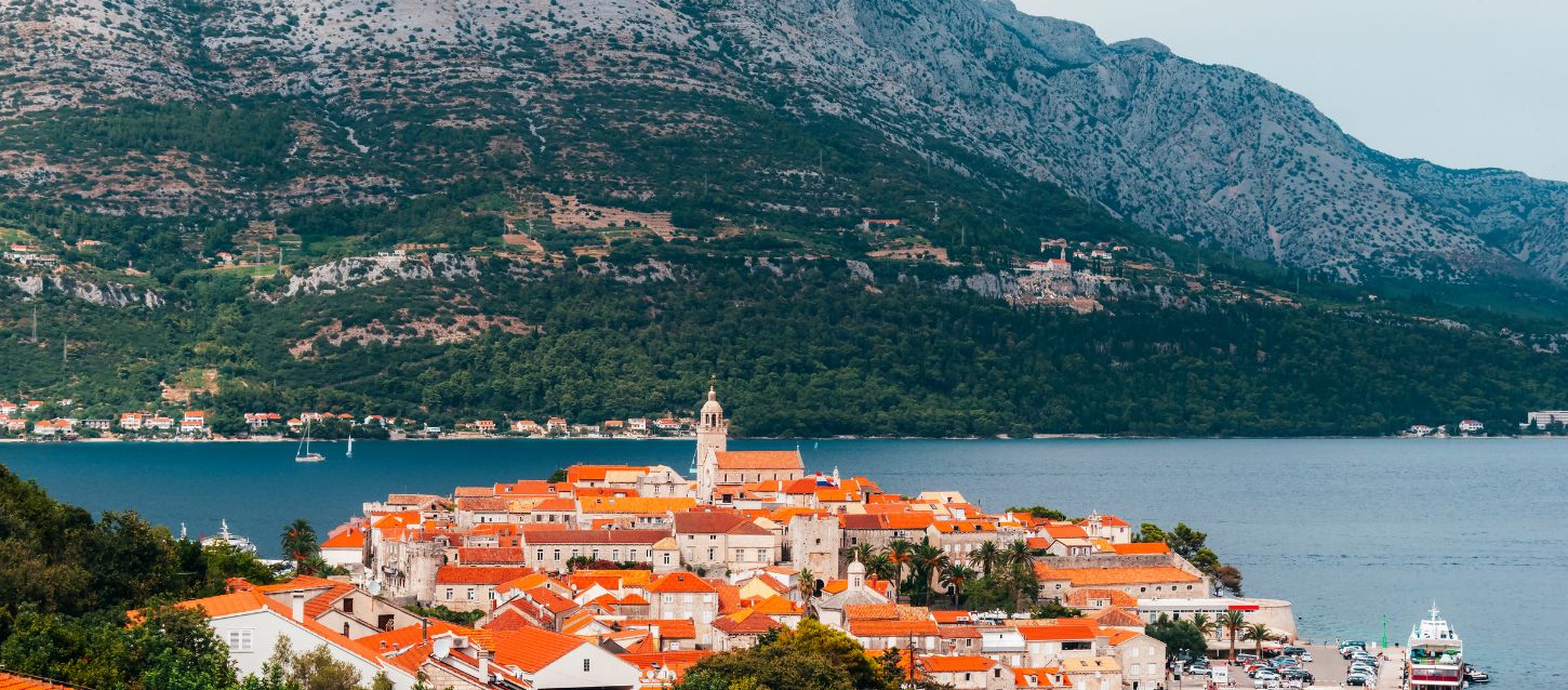 scenic view of the coastal town of Korčula, Croatia. The town, characterized by its red-tiled roofs and a prominent church tower, is situated on a small peninsula surrounded by the Adriatic Sea. In the background, a large, forested mountain rises steeply, creating a dramatic contrast with the calm blue water and the historic architecture of the town