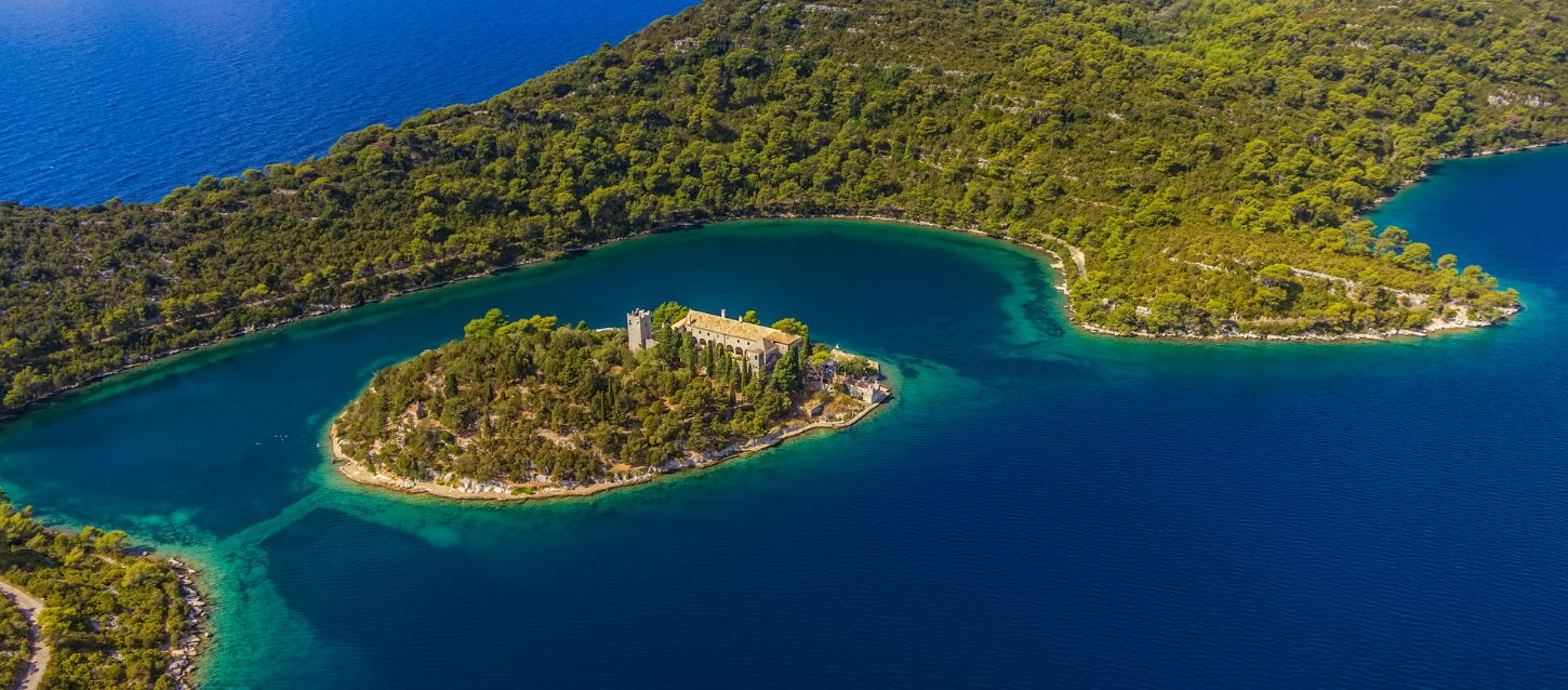 aerial view of a small, lush green island with a large building on it, situated in a body of deep blue water. The island is covered in dense forest, and the building appears to be a historic monastery or a similar structure with a prominent bell tower. The water surrounding the island is a vibrant turquoise near the shore, transitioning to a dark blue further out. The island is located within a larger, forested landmass, suggesting it is in a lake or a calm bay.
