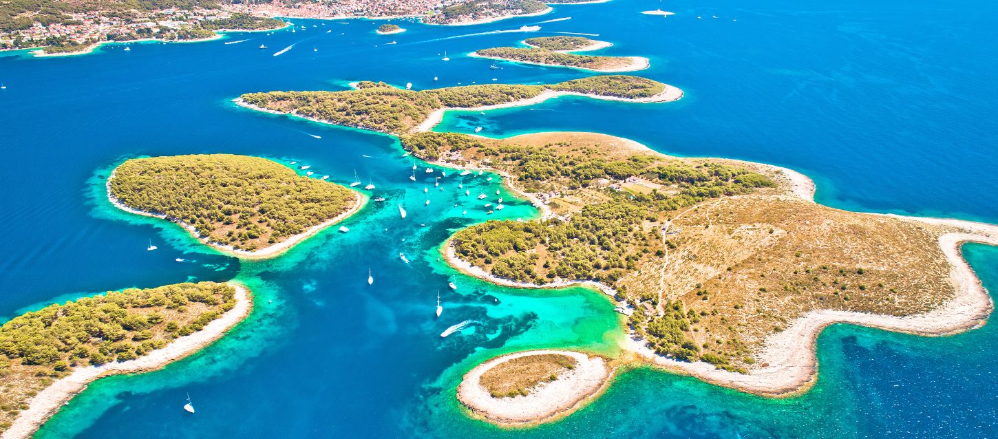 Aerial view of the Pakleni Islands, an archipelago located in the Adriatic Sea near the island of Hvar in Croatia. The image shows several small, lush green islands surrounded by incredibly clear turquoise and deep blue water. The islands are covered in dense vegetation, with some small, rocky coves and beaches visible along their irregular coastlines. The water is so transparent that the sandy seabed is visible in the shallow areas close to the shore. Several yachts sailing and at anchor are visible