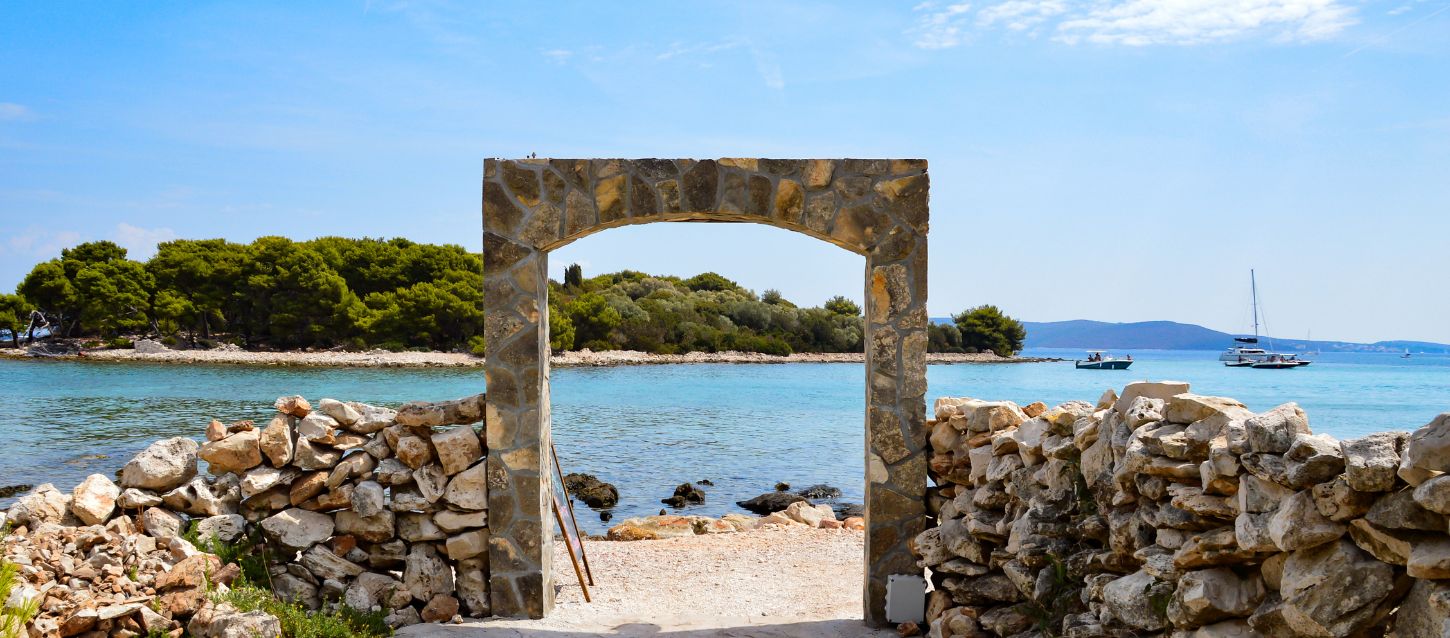 Stone archway and a low stone wall in the foreground, with a view of the sea and a small, forested island in the background. The water is turquoise, and the sky is blue with some clouds. Two boats are visible on the water