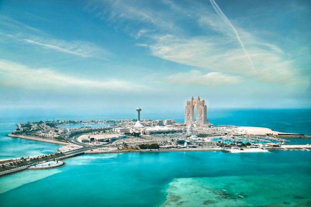 Aerial view of Abu Dhabi’s Corniche area, showing turquoise Gulf waters, modern skyscrapers, and a tall observation tower under a clear blue sky.
