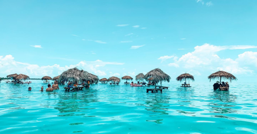 People relaxing in turquoise waters at Ambergris Caye, Belize, surrounded by wooden tables with thatched palm umbrellas.