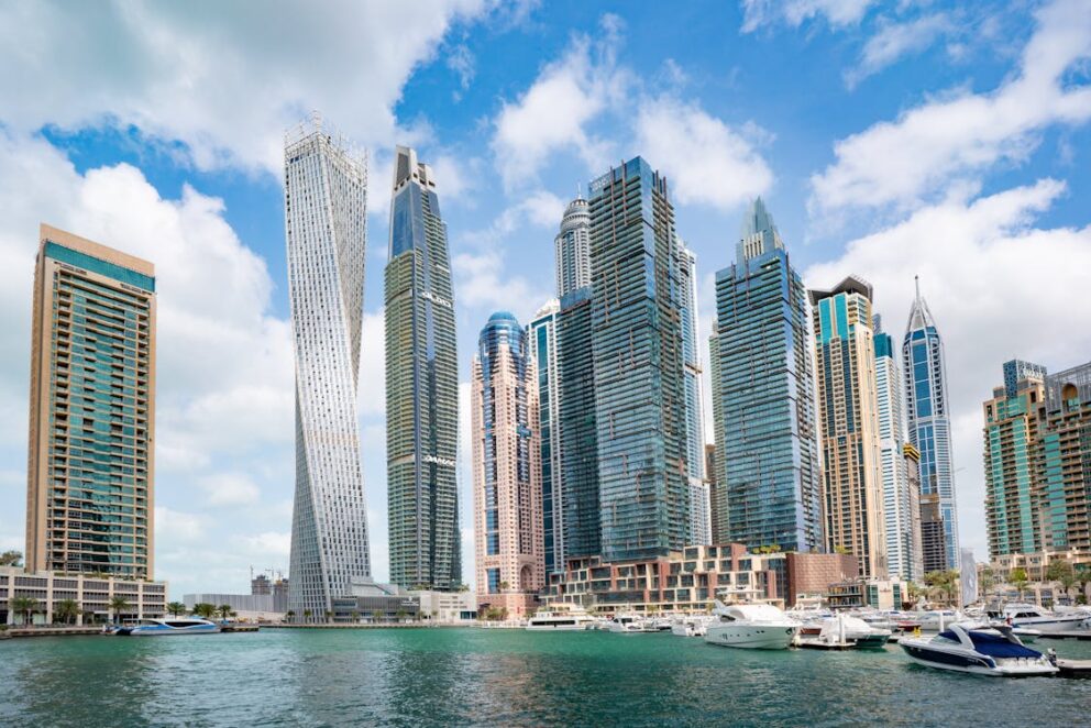 View of Dubai Marina with towering modern skyscrapers, luxury yachts docked along turquoise waters, and a partly cloudy blue sky above.