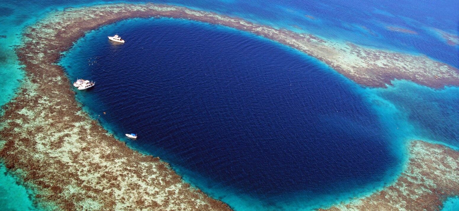 Aerial view of the Great Blue Hole in Belize, a deep circular sinkhole surrounded by turquoise coral reef and boats floating nearby.