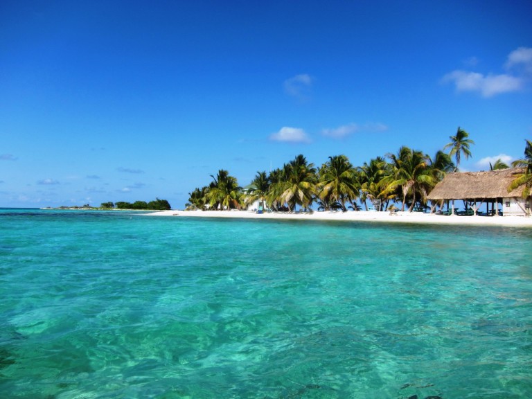 Turquoise waters and white sandy beach lined with palm trees and a thatched-roof hut at Placencia Beach, Belize, under a bright blue sky.
