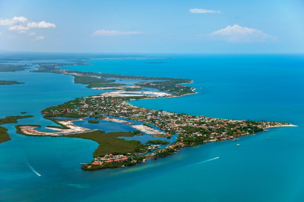 Aerial view of Placencia, Belize, showing a narrow peninsula surrounded by turquoise waters, dotted with houses, boats, and lush greenery.
