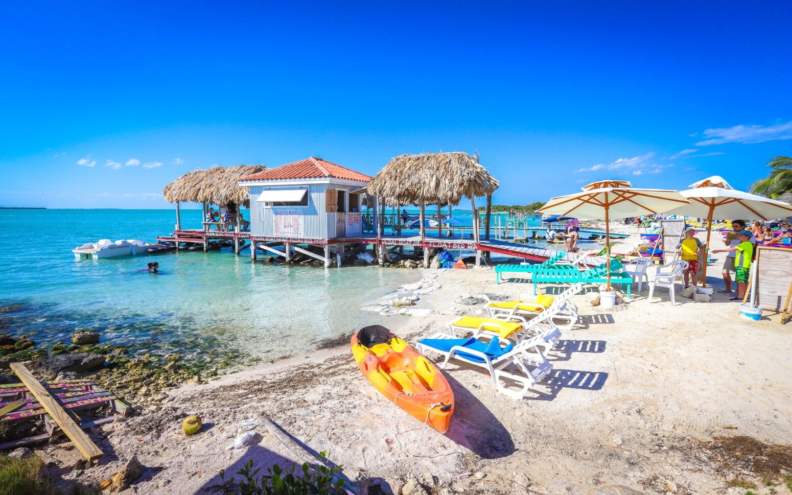 Colorful tropical beach scene at Secret Beach on Ambergris Caye, Belize, featuring lounge chairs, kayaks, umbrellas, and a small overwater bar with thatched roofs surrounded by clear turquoise water under a bright blue sky.