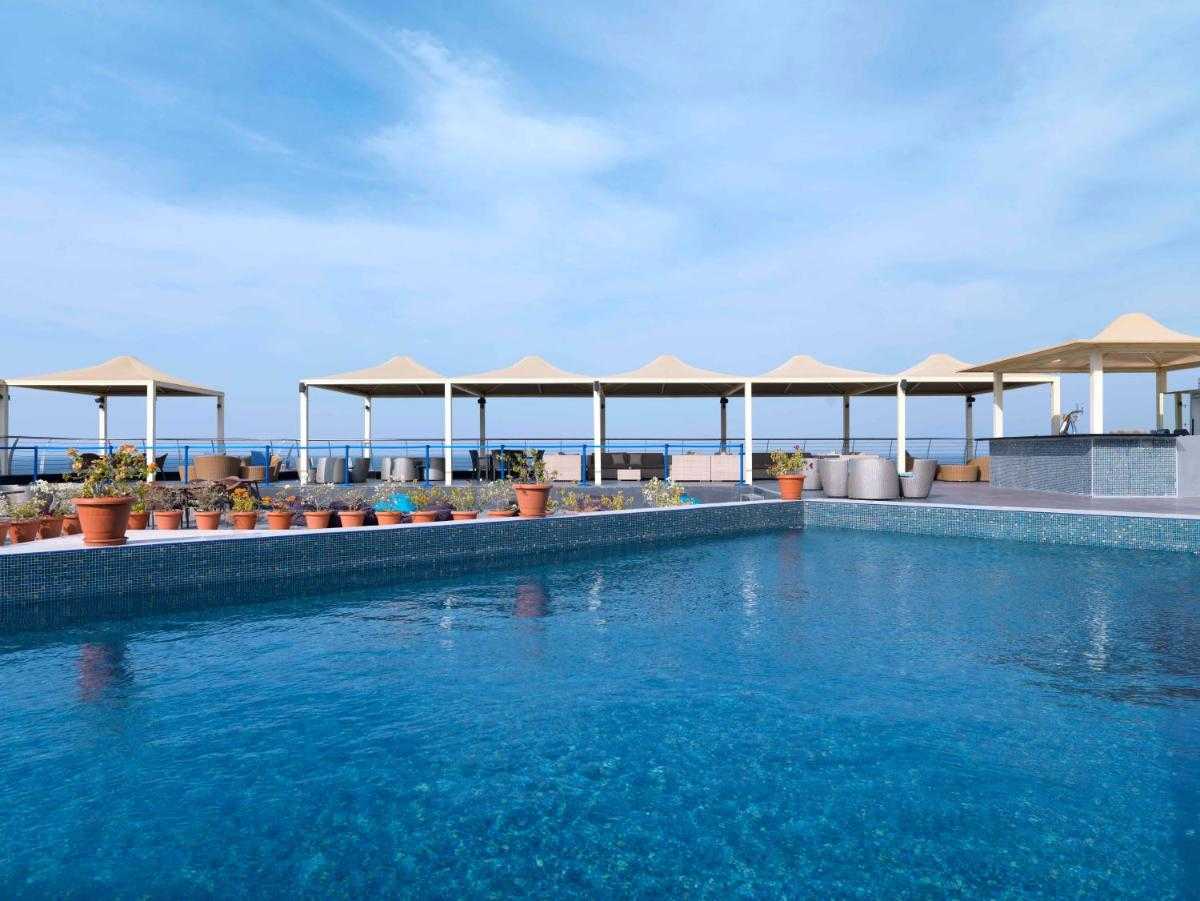 Rooftop pool at The Corniche in Muscat with clear blue water, terracotta potted plants, and shaded lounge areas under cream-colored canopies. The ocean and horizon are visible in the background beneath a bright blue sky.