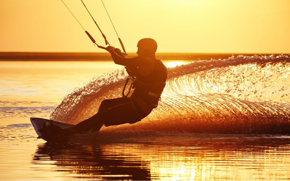 Person kite surfing on calm water at sunset, with golden light reflecting off the surface and a wave curling behind them in a dramatic spray.