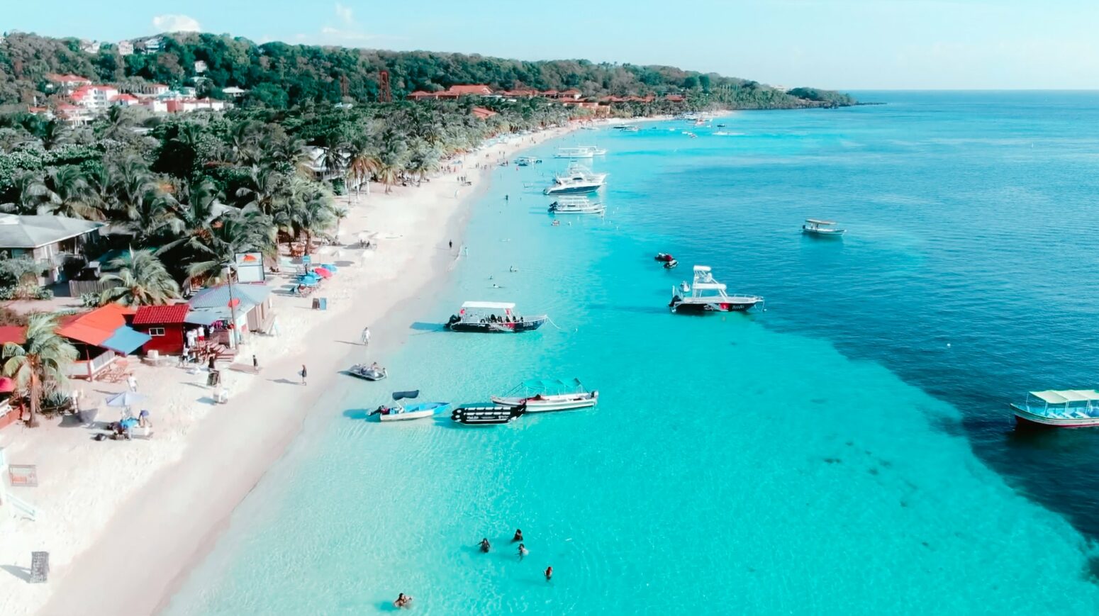 Aerial view of West Bay Beach in Roatán, Honduras, with turquoise water, white sand, palm trees, and boats anchored along the shoreline.