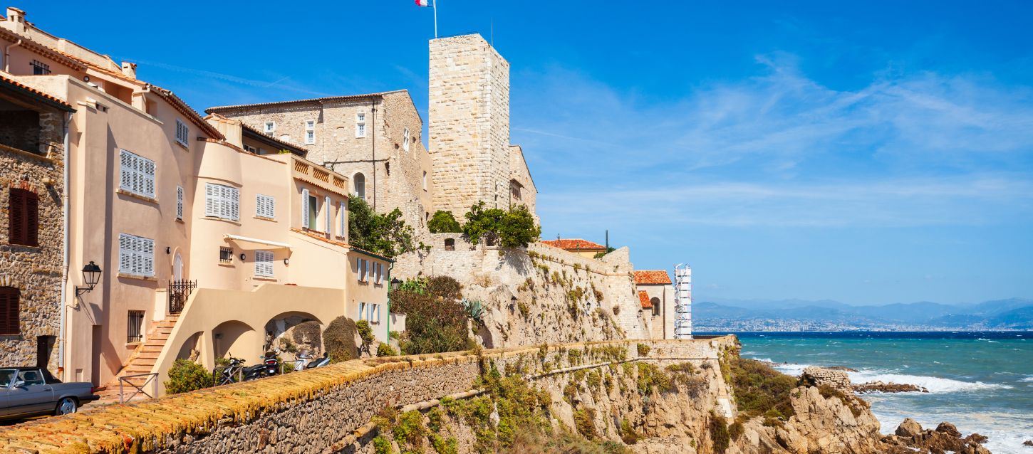 Scenic view of Antibes on the French Riviera featuring the Picasso Museum, old stone buildings, and fort walls overlooking the blue Mediterranean Sea under a clear sky.
