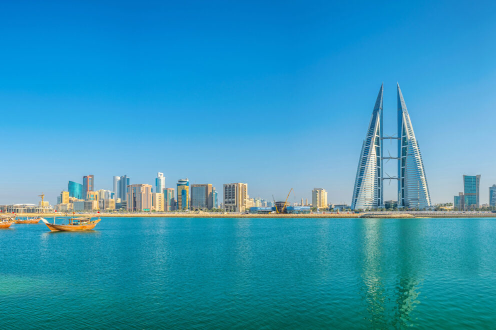 Modern city skyline with sleek skyscrapers and twin sail-shaped towers reflecting on calm turquoise waters, with traditional wooden boats floating in the foreground under a clear blue sky.