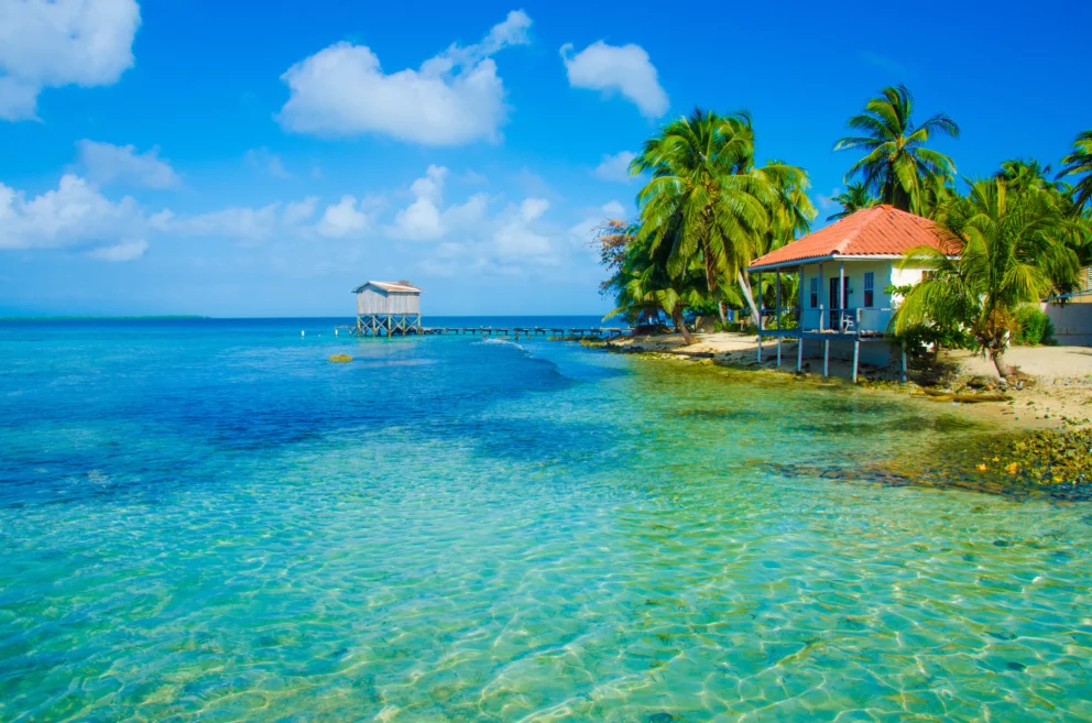 Tropical beach in Belize with clear turquoise water, palm trees, and a small house with a red roof on the shoreline.