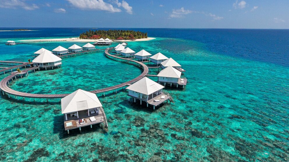Luxurious overwater villas connected by winding wooden walkways above vibrant turquoise waters, with a small tropical island and white sandy beach in the distance under a clear blue sky.